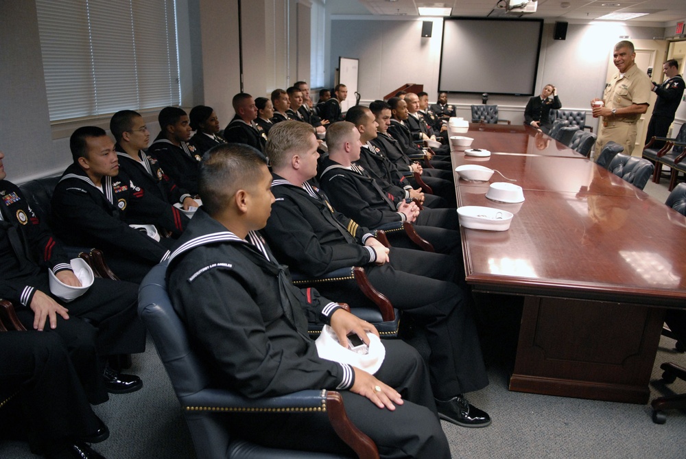 A group of U.S. Navy personnel in dress uniforms are seated in a formal conference room, listening to a senior officer or official who is standing and addressing them. The setting appears to be a briefing or orientation session, with a large polished conference table running along one side. White sailor caps (dixie cups) are held by several of the enlisted sailors.