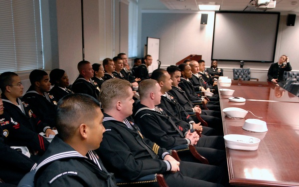 A group of U.S. Navy personnel in dress uniforms are seated in a formal conference room, listening to a senior officer or official who is standing and addressing them. The setting appears to be a briefing or orientation session, with a large polished conference table running along one side. White sailor caps (dixie cups) are held by several of the enlisted sailors.