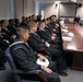A group of U.S. Navy personnel in dress uniforms are seated in a formal conference room, listening to a senior officer or official who is standing and addressing them. The setting appears to be a briefing or orientation session, with a large polished conference table running along one side. White sailor caps (dixie cups) are held by several of the enlisted sailors.