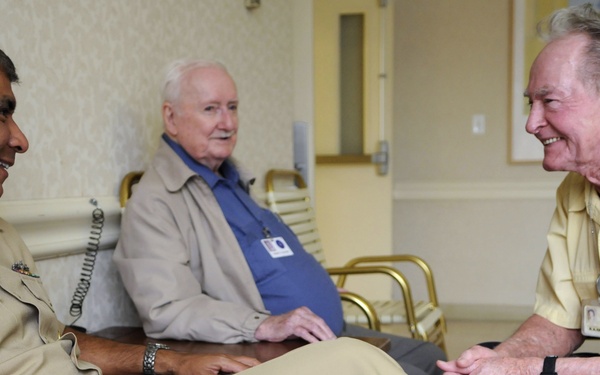 A U.S. Navy enlisted service member in tan/khaki uniform is seated and engaged in conversation with two elderly civilian men, likely in a care facility or veterans' home setting. The interaction appears friendly and positive, with all three individuals smiling. This appears to be a military outreach or veteran visitation event.