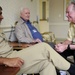 A U.S. Navy enlisted service member in tan/khaki uniform is seated and engaged in conversation with two elderly civilian men, likely in a care facility or veterans' home setting. The interaction appears friendly and positive, with all three individuals smiling. This appears to be a military outreach or veteran visitation event.