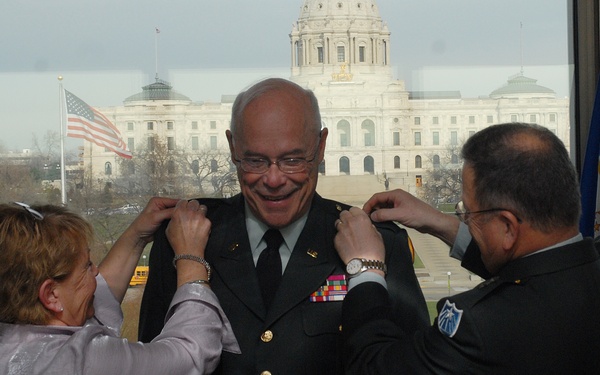 A military promotion or pinning ceremony is taking place indoors with a large capitol building visible through the window behind the subjects. A woman on the left and a uniformed military officer on the right are simultaneously pinning rank insignia onto the collar of a smiling man in Army dress green uniform. An American flag and what appears to be a state or military flag flank the scene.