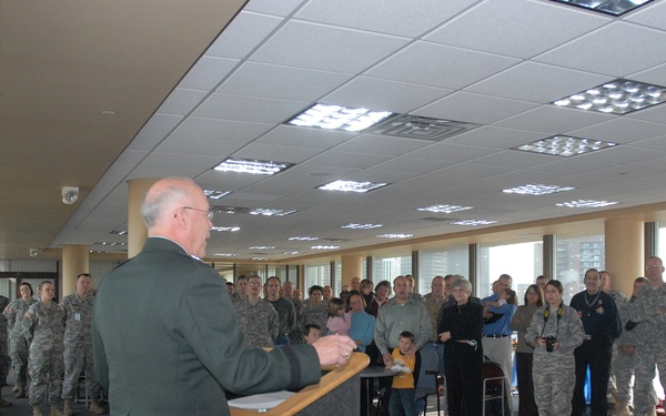 A military official in dress greens is addressing a large group of uniformed soldiers and civilians from a podium in what appears to be a modern indoor facility with large windows. The audience consists predominantly of personnel in Army Combat Uniforms (ACUs) along with some civilians, including children. The setting suggests a formal military briefing, ceremony, or unit gathering.