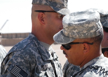 The image shows multiple U.S. Army soldiers in Army Combat Uniform (ACU) digital camouflage pattern. A soldier with the name tape 'PEREZ' is receiving what appears to be a medal or award being pinned to his uniform by another soldier with the partial name tape 'ALEX...' visible. The setting appears to be an outdoor military environment in an arid or desert location.