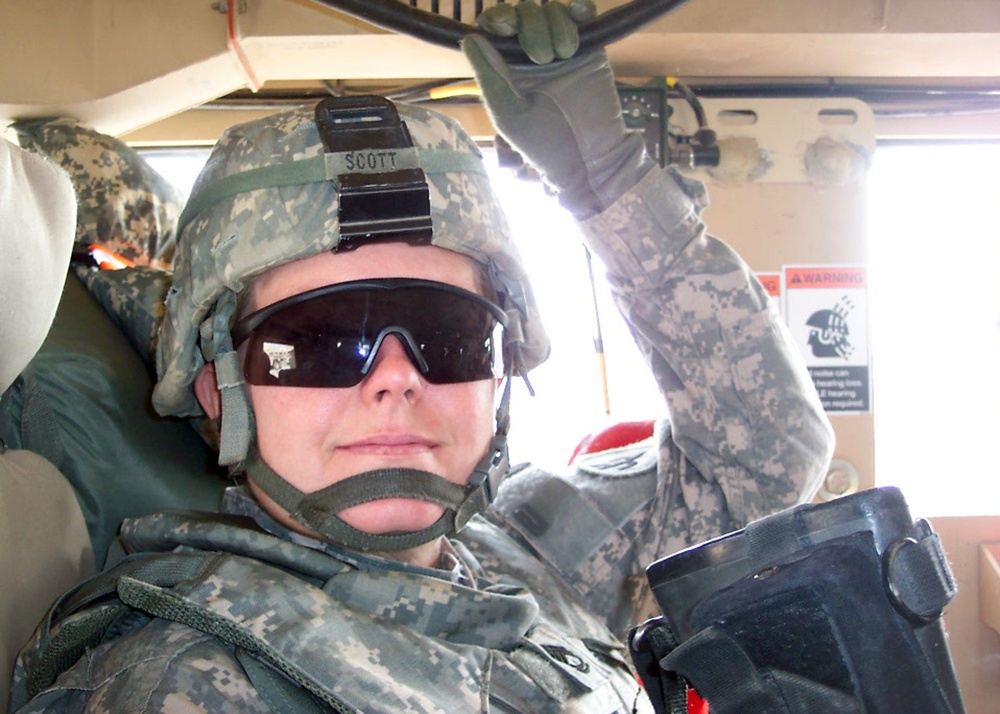 A U.S. Army soldier with the last name 'Scott' is seated inside a military vehicle, likely a Humvee. The soldier is wearing ACU (Army Combat Uniform) digital camouflage, a combat helmet with NVG mount, ballistic eyewear, tactical gloves, and body armor. One hand is raised, possibly adjusting equipment or operating a vehicle hatch mechanism.