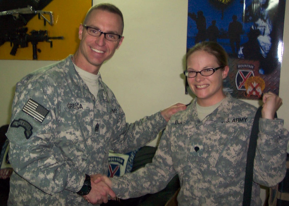 Two U.S. Army soldiers in ACU (Army Combat Uniform) digital camouflage are shaking hands and smiling for a photo. The soldier on the left has the name tape 'GRECA' and wears a Ranger tab and 10th Mountain Division patch. The setting appears to be an indoor military office or common area.