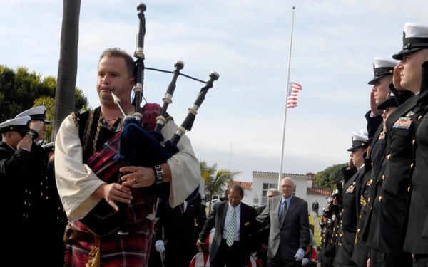 A military funeral procession is taking place outdoors, led by a bagpiper dressed in traditional Scottish Highland attire including a tartan kilt. Uniformed military personnel, appearing to be U.S. Navy officers, line both sides of a walkway in formal dress uniforms standing at attention or saluting. A flag-draped casket is visible being carried in the background, with an American flag flying at half-staff.