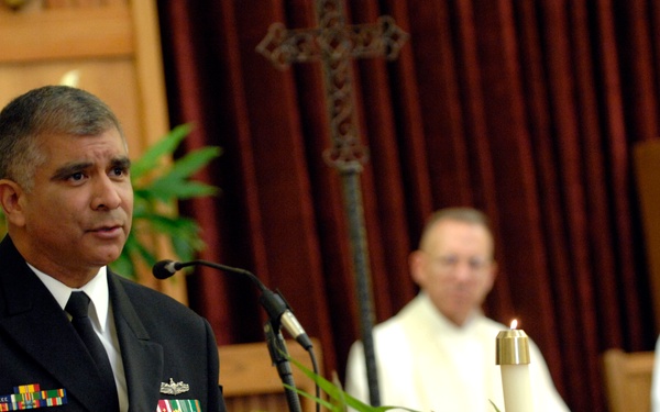 A military officer in dress uniform with multiple medals and ribbons speaks at a wooden podium equipped with a microphone, inside what appears to be a chapel or church setting. Two clergy members dressed in white vestments are seated in the background. The setting suggests a formal religious or memorial ceremony.