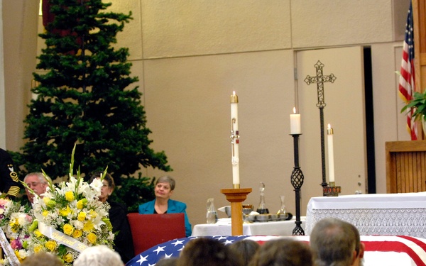 A military funeral service is taking place inside a church or chapel. A flag-draped casket is positioned at the front of the room, with a uniformed military officer delivering remarks at a podium. A portrait photograph of the deceased in military uniform is displayed to the right of the casket.