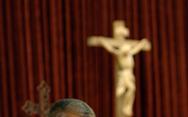 A military officer in a dark dress uniform adorned with multiple medals and ribbon bars stands at a wooden podium with a microphone, appearing to speak. The setting is a Catholic church, evidenced by a prominent crucifix on a wooden stand and a decorative cross on the wall behind red curtains. A second individual dressed in white liturgical vestments is seated in the background.