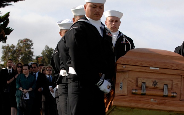A U.S. Navy military funeral is taking place at a national cemetery. Navy pallbearers in dress uniforms are carrying a wooden casket while mourners observe in the background. A visible headstone identifies the grave of Thomas S. Crow, 4th Master Chief Petty Officer of the Navy (MCPON), Vietnam veteran, who lived from March 16, 1934 to November 30, 2008.