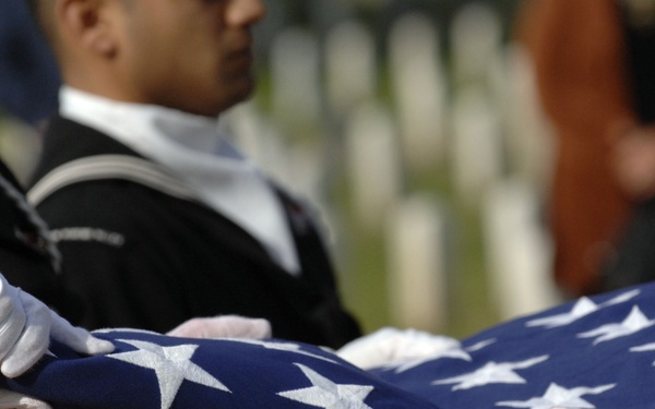 The image depicts a military funeral flag-folding ceremony, with uniformed service members in white gloves carefully folding an American flag. The blue field with white stars is prominently visible as multiple hands handle the flag with precision. White grave markers are visible in the blurred background, confirming a military cemetery setting.