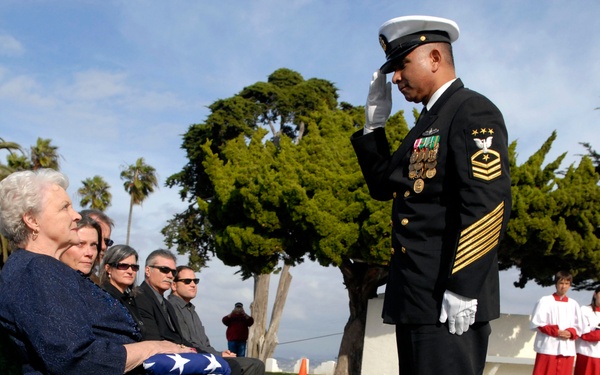 A military funeral ceremony is taking place outdoors, likely at a national cemetery. A senior Navy enlisted servicemember in dress uniform is rendering a salute to a seated elderly woman who holds a folded American flag on her lap. Mourners, clergy in red robes, and other attendees are visible in the background.