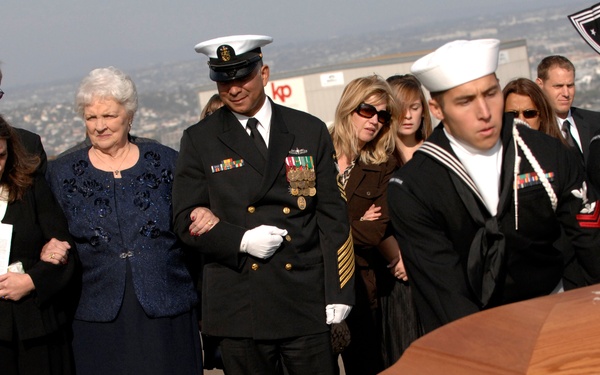 A military funeral ceremony is taking place at what appears to be a hillside cemetery with a city visible in the background. U.S. Navy sailors in dress uniforms are serving as pallbearers, carrying a wooden casket while grieving family members look on. A senior Navy officer in dress blues with medals is escorting an elderly woman, while other mourners dressed in black show visible signs of grief.