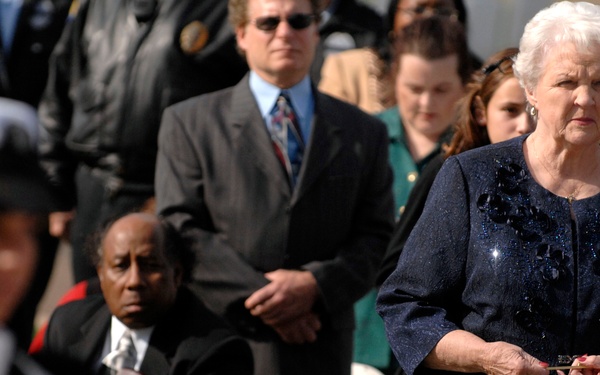 A solemn military funeral or memorial ceremony is taking place outdoors. A Navy service member in dress whites and white gloves is folding or presenting an American flag in the foreground, while a group of civilians, including an elderly woman in a dark navy suit, observe with serious expressions. The scene suggests a formal military honors ceremony, likely at a burial or memorial service.