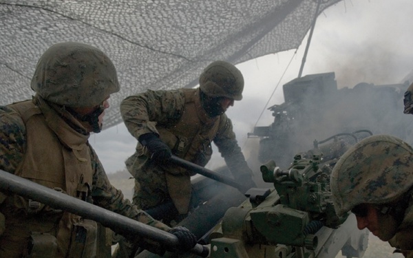 Multiple military personnel in digital camouflage uniforms are operating an artillery piece under a camouflage net. Smoke is visible around the weapon system, suggesting it has recently been fired or is in the process of being operated. The soldiers appear to be performing coordinated crew-served weapon duties.