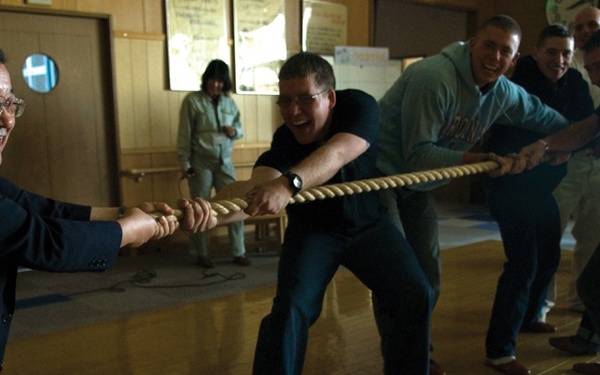A group of people are engaged in an indoor tug-of-war competition, pulling a thick rope in opposite directions. On the left side, a man in a dark business suit and glasses is straining with effort, while a line of casually dressed individuals pulls from the right. The activity appears to be taking place in a gymnasium or community hall with wooden flooring.