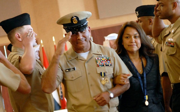 A military ceremony appears to be taking place, likely a retirement or promotion event for a senior U.S. Navy officer. The central figure, a high-ranking Navy Chief Petty Officer or Master Chief wearing dress khaki uniform with multiple ribbons and medals, is escorted by a civilian woman through a corridor of saluting sailors. The surrounding personnel are rendering honors by saluting as the pair passes.