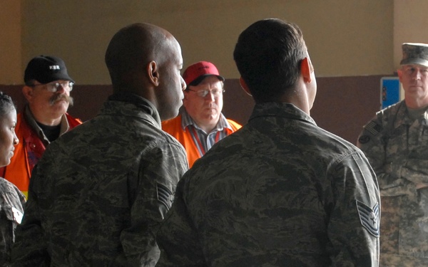 A group of military personnel in camouflage uniforms are gathered in what appears to be a large indoor facility, possibly a hangar or warehouse. Several civilians in orange safety vests and casual clothing are also present among the group. The scene suggests a briefing, coordination meeting, or joint civil-military operation.