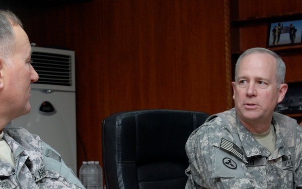 Two U.S. Army soldiers in ACU (Army Combat Uniform) camouflage are seated at a wooden conference table engaged in conversation. The soldier on the left wears a 101st Airborne Division patch, while the soldier on the right appears to be gesturing as he speaks. A third soldier is partially visible in the background.