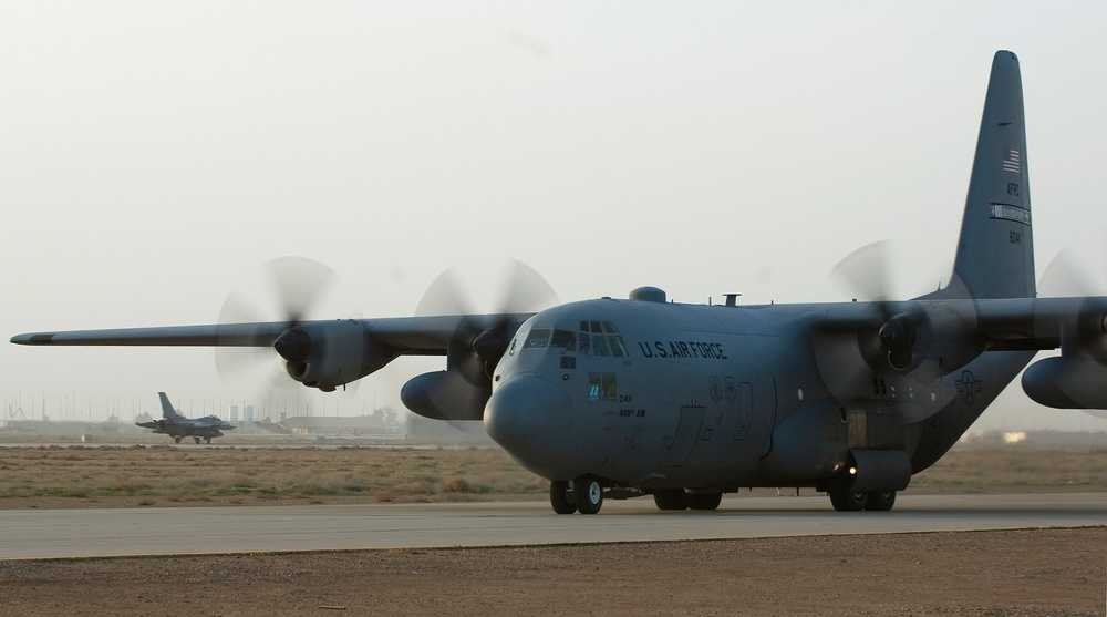 A U.S. Air Force C-130 Hercules transport aircraft is taxiing on a runway at what appears to be a desert airbase, with all four turboprop engines running and propellers in motion. In the background, a fighter jet (likely an F-16) is also visible on the tarmac. The environment suggests an arid, Middle Eastern or Southwest Asian operating location.