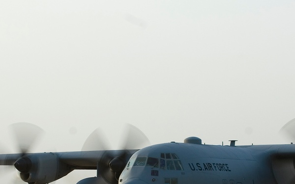 A U.S. Air Force C-130 Hercules transport aircraft is taxiing on a runway at what appears to be a desert airbase, with all four turboprop engines running and propellers in motion. In the background, a fighter jet (likely an F-16) is also visible on the tarmac. The environment suggests an arid, Middle Eastern or Southwest Asian operating location.