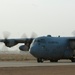 A U.S. Air Force C-130 Hercules transport aircraft is taxiing on a runway at what appears to be a desert airbase, with all four turboprop engines running and propellers in motion. In the background, a fighter jet (likely an F-16) is also visible on the tarmac. The environment suggests an arid, Middle Eastern or Southwest Asian operating location.