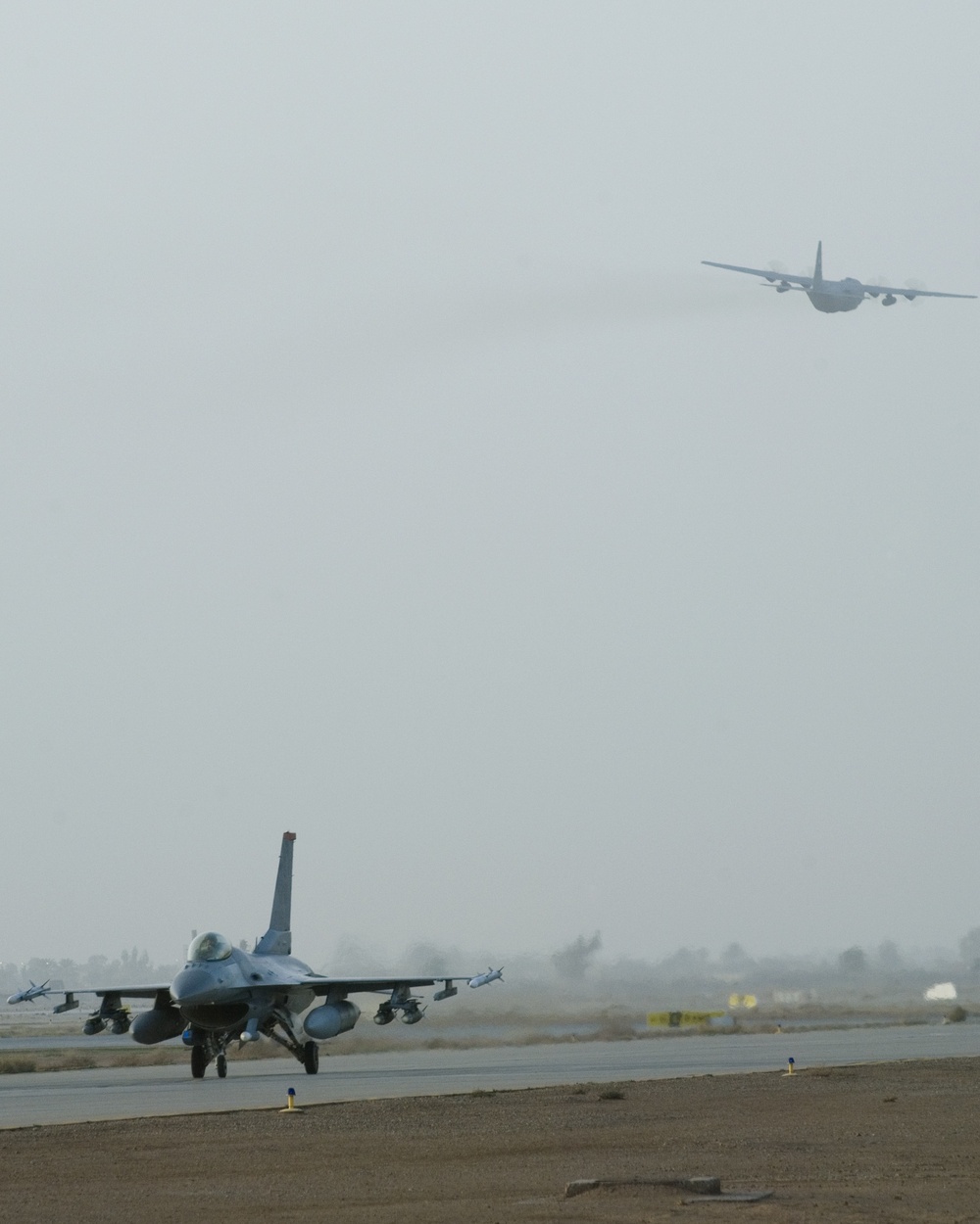 A military airfield scene showing an F-16 Fighting Falcon jet fighter taxiing on a runway in the foreground, while a large four-engine transport aircraft (consistent with a C-130 Hercules) flies overhead in hazy conditions. The environment appears arid and dusty, suggesting a forward operating base in a desert region. The overcast, low-visibility sky reduces overall image clarity.