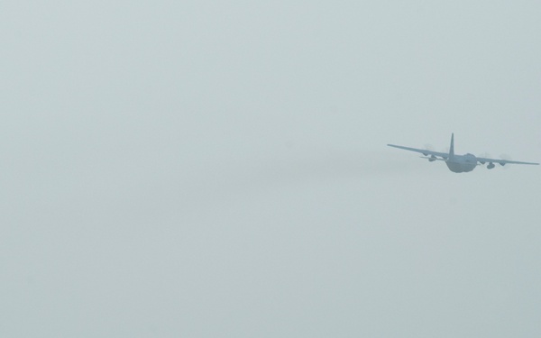 A military airfield scene showing an F-16 Fighting Falcon jet fighter taxiing on a runway in the foreground, while a large four-engine transport aircraft (consistent with a C-130 Hercules) flies overhead in hazy conditions. The environment appears arid and dusty, suggesting a forward operating base in a desert region. The overcast, low-visibility sky reduces overall image clarity.