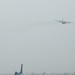 A military airfield scene showing an F-16 Fighting Falcon jet fighter taxiing on a runway in the foreground, while a large four-engine transport aircraft (consistent with a C-130 Hercules) flies overhead in hazy conditions. The environment appears arid and dusty, suggesting a forward operating base in a desert region. The overcast, low-visibility sky reduces overall image clarity.