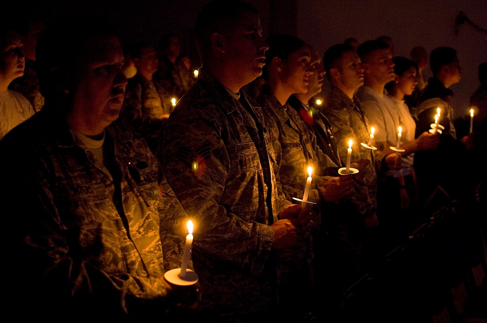 A group of military personnel in camouflage uniforms are gathered in a darkened space, each holding lit candles during what appears to be a candlelight vigil or memorial ceremony. The warm glow of the candles illuminates their faces as they stand in rows, some appearing to sing or recite something. The solemn atmosphere and uniformed participants suggest a formal military commemorative event.