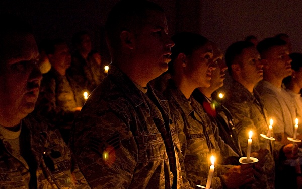 A group of military personnel in camouflage uniforms are gathered in a darkened space, each holding lit candles during what appears to be a candlelight vigil or memorial ceremony. The warm glow of the candles illuminates their faces as they stand in rows, some appearing to sing or recite something. The solemn atmosphere and uniformed participants suggest a formal military commemorative event.