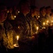 A group of military personnel in camouflage uniforms are gathered in a darkened space, each holding lit candles during what appears to be a candlelight vigil or memorial ceremony. The warm glow of the candles illuminates their faces as they stand in rows, some appearing to sing or recite something. The solemn atmosphere and uniformed participants suggest a formal military commemorative event.