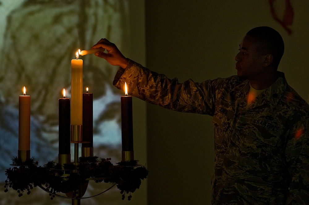 A U.S. Air Force service member in ABU (Airman Battle Uniform) camouflage is lighting candles on what appears to be an Advent wreath candelabra. The scene is dimly lit, with the candlelight providing warm illumination against a dark background. A projected image is visible on the wall behind the subject.