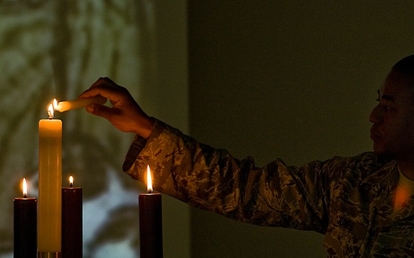 A U.S. Air Force service member in ABU (Airman Battle Uniform) camouflage is lighting candles on what appears to be an Advent wreath candelabra. The scene is dimly lit, with the candlelight providing warm illumination against a dark background. A projected image is visible on the wall behind the subject.
