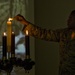 A U.S. Air Force service member in ABU (Airman Battle Uniform) camouflage is lighting candles on what appears to be an Advent wreath candelabra. The scene is dimly lit, with the candlelight providing warm illumination against a dark background. A projected image is visible on the wall behind the subject.