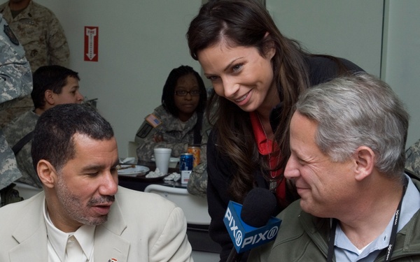 A female reporter from PIX11 news is conducting an interview with two men seated at a table, apparently at a military dining facility or event. Several people in U.S. Army camouflage uniforms are visible in the background. The setting appears to be an informal meal or gathering with food items visible on the table.