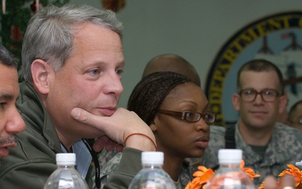 A group of civilians and military personnel are seated together at a dining table in what appears to be a Navy facility, indicated by the 'Department of the Navy' seal visible on the wall in the background. The gathering appears to be an official meeting or luncheon, with water bottles, condiments, flowers, and cups arranged on the table. The mix of civilian attire and military camouflage uniforms suggests a formal visit or briefing event.