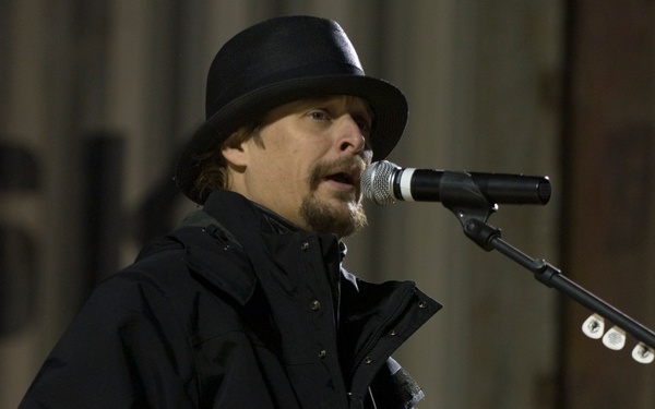 A male performer is on stage singing into a microphone while playing an electric guitar. He is dressed in all black, including a black jacket and a black wide-brimmed fedora-style hat, and has a goatee. The image appears to be taken at a live concert or outdoor performance event.