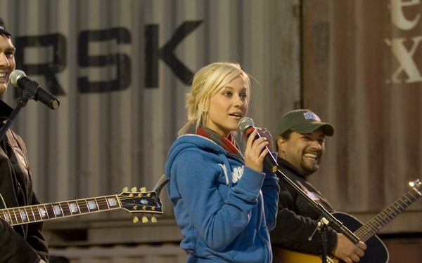 Three performers are on an outdoor stage in front of what appears to be shipping containers with partial text 'ARSK' visible (likely Maersk). A blonde woman in a blue hoodie holds a microphone and sings, flanked by two men seated on stools playing acoustic guitars. The setting appears to be an informal or military-style venue performance.