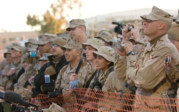 A large group of military personnel in desert camouflage uniforms are gathered behind orange safety netting, watching an off-camera event. Several individuals are photographing or filming the event using cameras and video equipment. The setting appears to be a military base or installation in an arid environment.