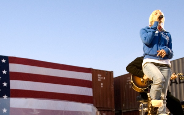 An outdoor concert performance appears to be taking place at a military base or deployment location, evidenced by the large American flag backdrop and shipping containers in the background. A blonde performer in a blue hoodie and boots stands elevated on a stage platform singing into a microphone, while a guitarist performs in the background. An audience is visible in the lower portion of the image, reflected in what appears to be a transparent barrier.