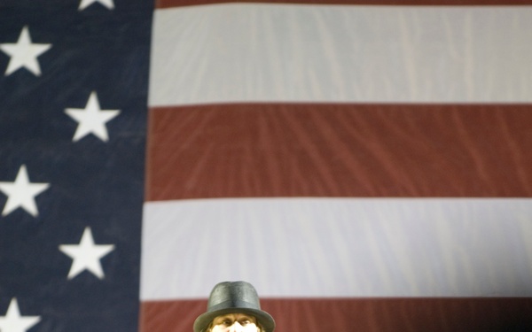 A male performer dressed in all black, wearing a dark fedora-style hat, is singing into a microphone while playing an electric guitar. He is performing in front of a large American flag backdrop, suggesting a patriotic or political event. The foreground shows silhouetted audience members watching the performance.