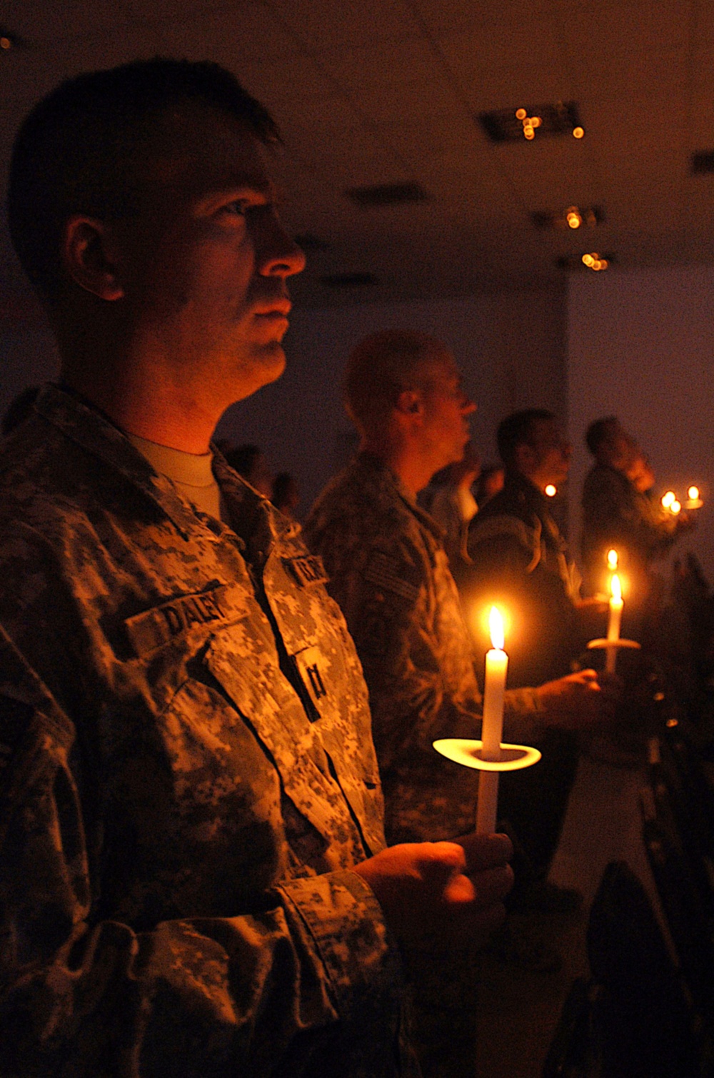 A group of military personnel in camouflage uniforms are participating in a candlelight ceremony in a darkened indoor venue. The foreground soldier, whose name tape reads 'DALEY', holds a lit candle on a small wax-catching holder. Multiple service members are visible in the background, each holding lit candles, suggesting a memorial or solemn commemorative event.