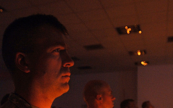 A group of military personnel in camouflage uniforms are participating in a candlelight ceremony in a darkened indoor venue. The foreground soldier, whose name tape reads 'DALEY', holds a lit candle on a small wax-catching holder. Multiple service members are visible in the background, each holding lit candles, suggesting a memorial or solemn commemorative event.