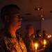 A group of military personnel in camouflage uniforms are participating in a candlelight ceremony in a darkened indoor venue. The foreground soldier, whose name tape reads 'DALEY', holds a lit candle on a small wax-catching holder. Multiple service members are visible in the background, each holding lit candles, suggesting a memorial or solemn commemorative event.
