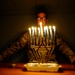 A U.S. Air Force service member in camouflage uniform sits behind a fully lit Hanukkah menorah (Chanukiah) with all nine candles burning. The image is dramatically lit primarily by the candlelight against a dark background, creating a striking ceremonial atmosphere. The menorah features a Star of David base design and is placed on a wooden table.