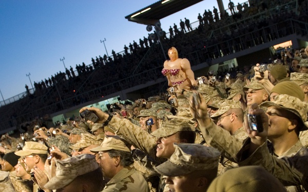 A large crowd of military personnel in desert camouflage uniforms are gathered in what appears to be a stadium or outdoor venue at dusk. The soldiers appear to be attending an entertainment or morale event, with many cheering, taking photos, and raising their hands. A novelty inflatable figure is visible being held up above the crowd.