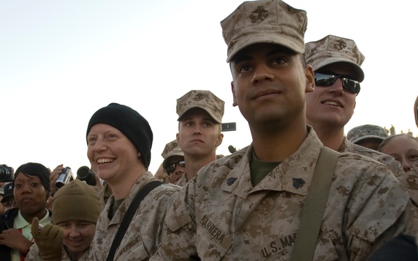 A group of U.S. Marines in desert camouflage (MARPAT) uniforms are gathered behind an orange safety barrier fence, appearing to watch an event or performance. Several civilians and military personnel are visible taking photographs with cameras. The mood appears festive and relaxed, suggesting a morale or entertainment event.