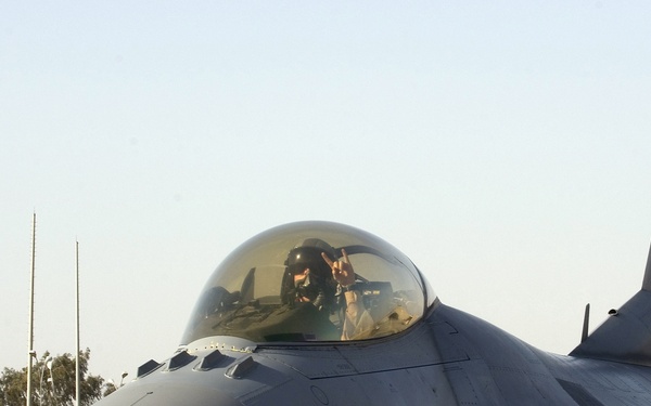 A U.S. Air Force F-16 Fighting Falcon fighter jet is photographed on a tarmac, likely at a forward operating base in a Middle Eastern location based on the arid environment and architecture visible in the background. The pilot is seated in the cockpit and is making a peace/victory hand gesture through the canopy. The aircraft bears the tail code 'WW' and serial number ending in '912', consistent with a unit based at Misawa Air Base, Japan.